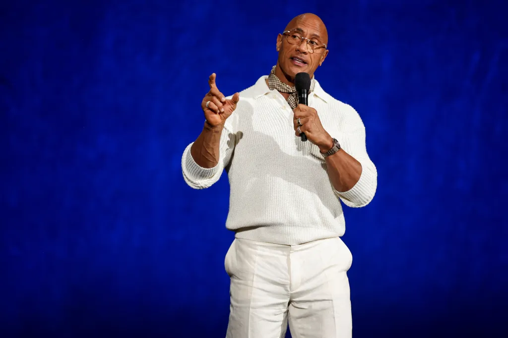 Dwayne Johnson, wearing a white sweater and pants with a patterned scarf, speaks into a microphone at CinemaCon.