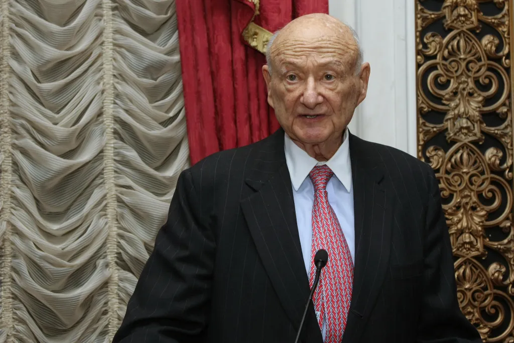 Ed Koch speaking at an event, wearing a suit and red tie, with a microphone in front of him.