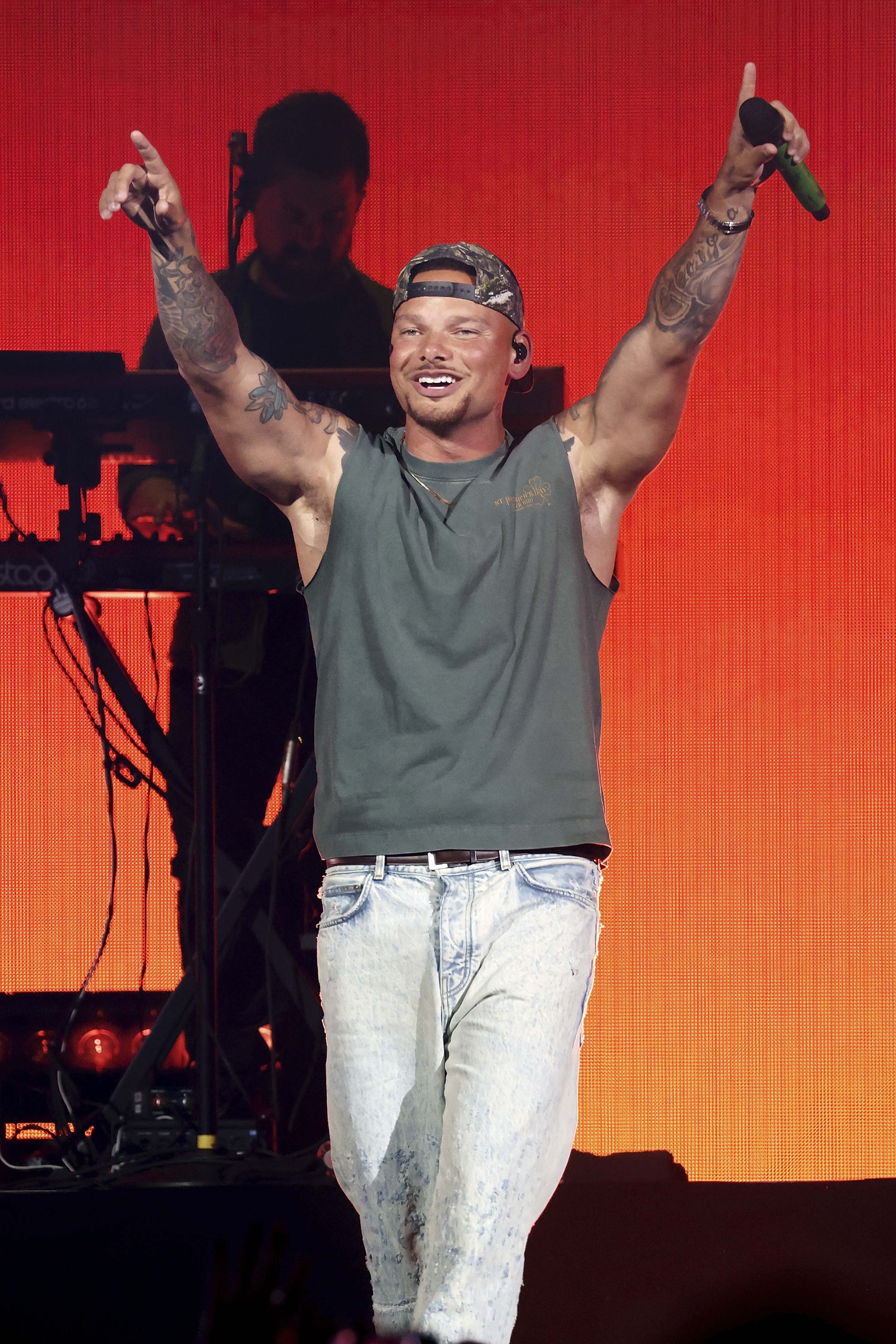 Kane Brown performing on stage with his arms raised, holding a microphone in his right hand.