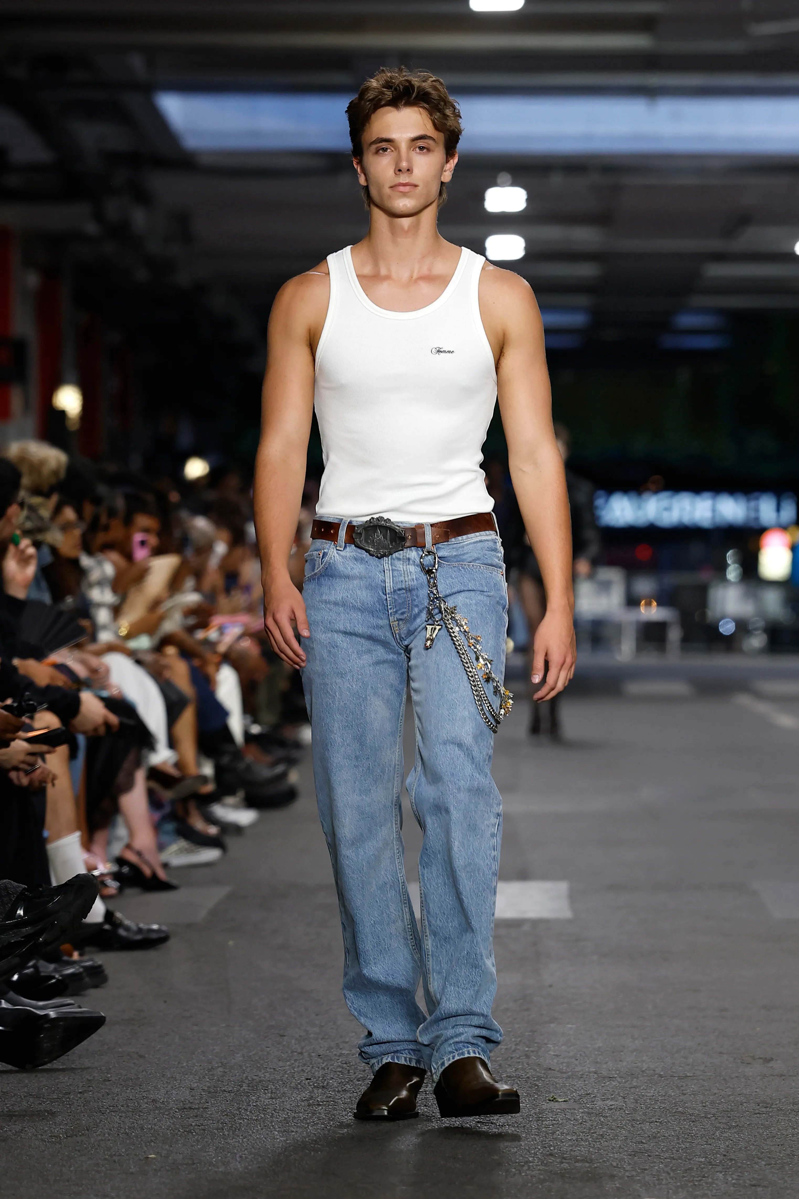 A model in a white tank top and light wash jeans with a decorative belt and chains walks the runway.