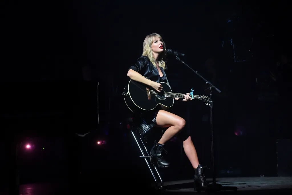 Taylor Swift performing on stage at the City of Lover concert, sitting on a stool and playing an acoustic guitar.