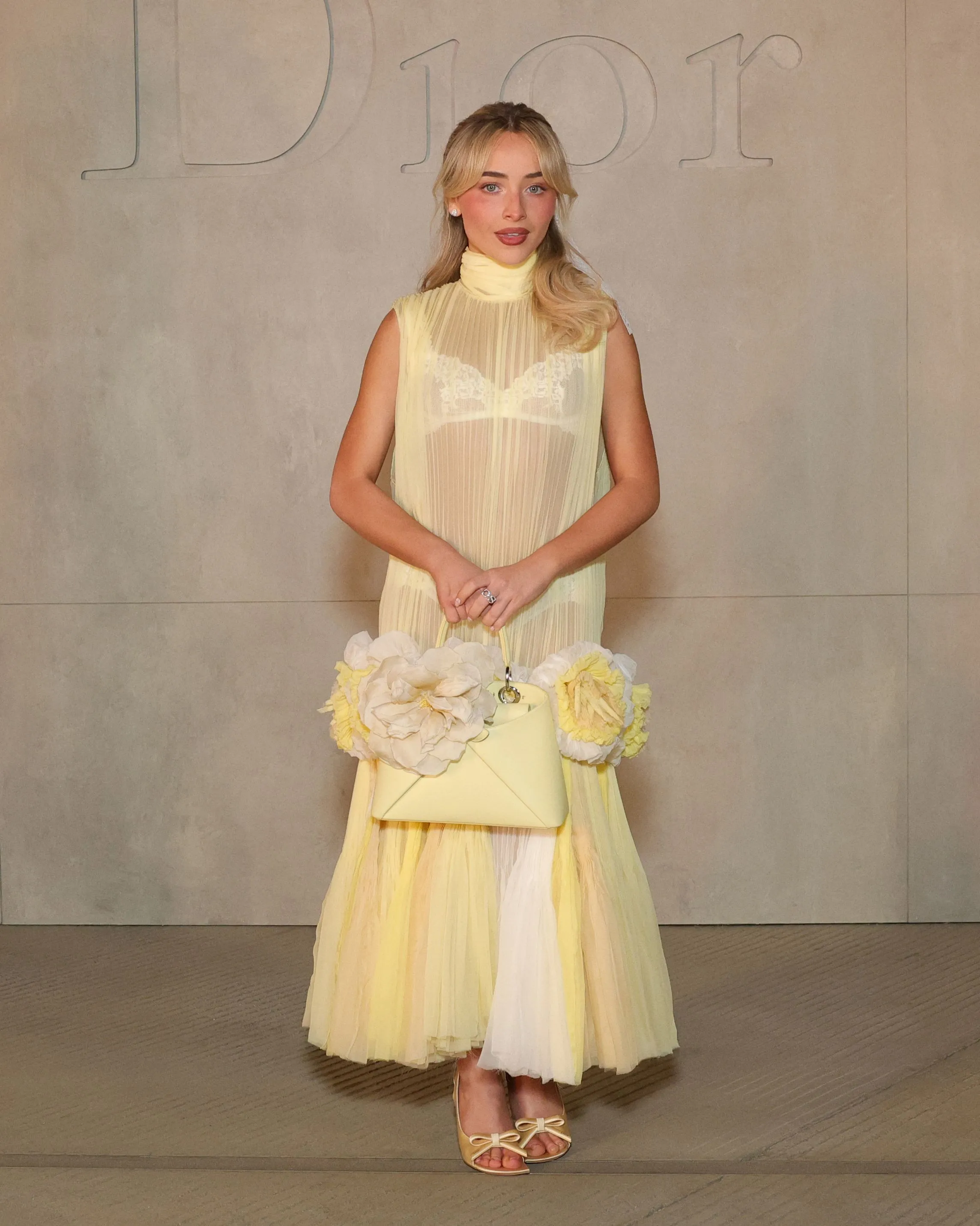 Sabrina Carpenter in a sheer yellow pleated dress with a matching flower-adorned purse.