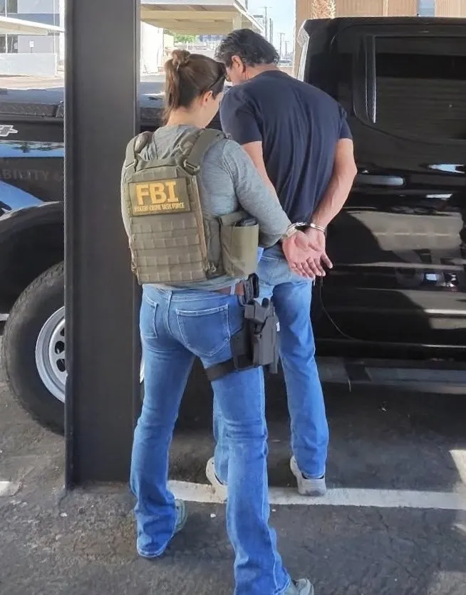 An FBI agent in tactical gear with a firearm holstered on her thigh apprehends a man in handcuffs.