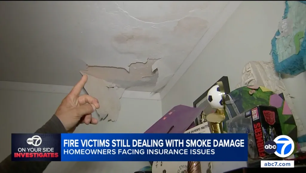 A person's finger pointing at a severely damaged ceiling with peeling paint and discoloration from smoke.