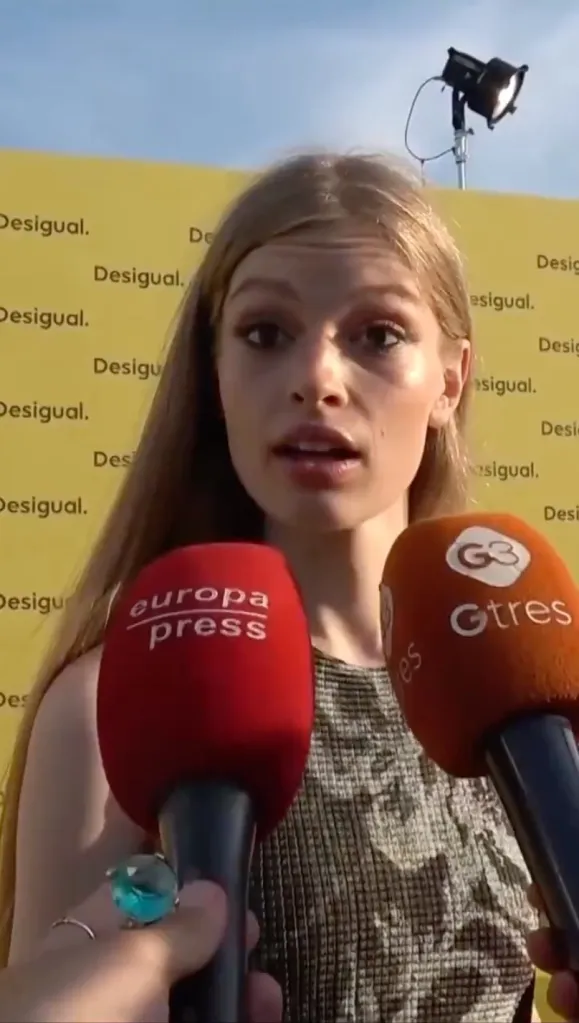 A young woman with long light brown hair is being interviewed by two microphones from Europa Press and Gtres, in front of a yellow banner with the word 