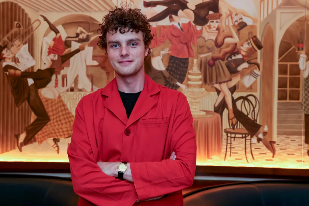 Actor Cameron Scott Roberts, in a red jacket, stands in front of a mural depicting dancing figures.