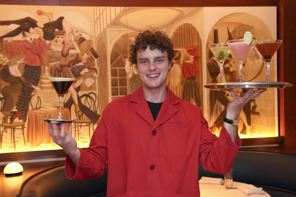 Actor Cameron Scott Roberts in a red jacket holding trays of cocktails.