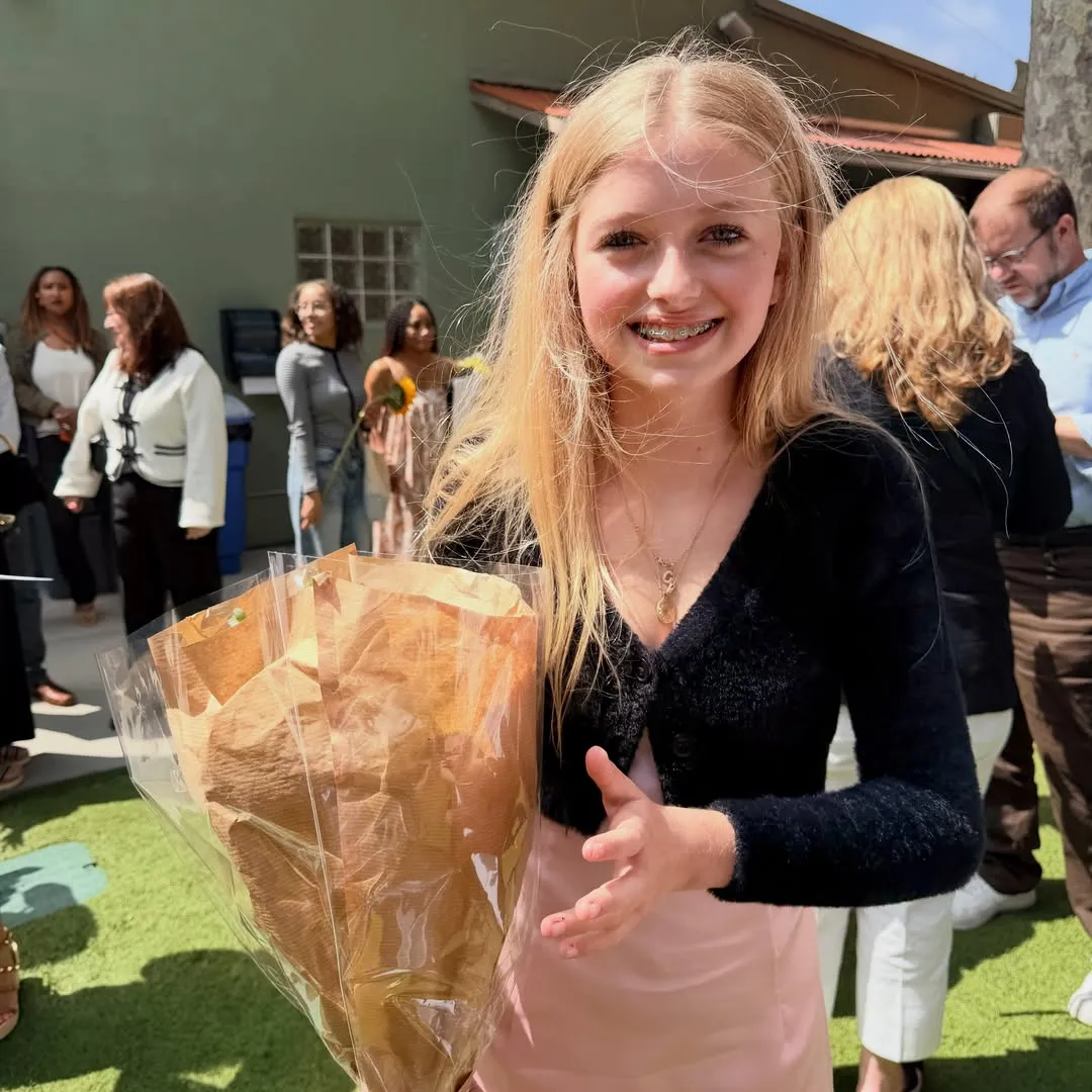 A young blonde woman wearing braces and a black cardigan over a pink dress smiles while holding a bouquet of flowers wrapped in brown paper and clear plastic.