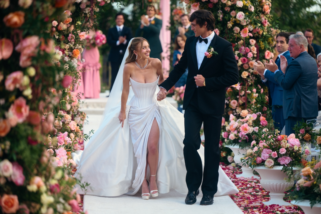 A bride in a white gown and a groom in a tuxedo walk through a floral archway as guests applaud.