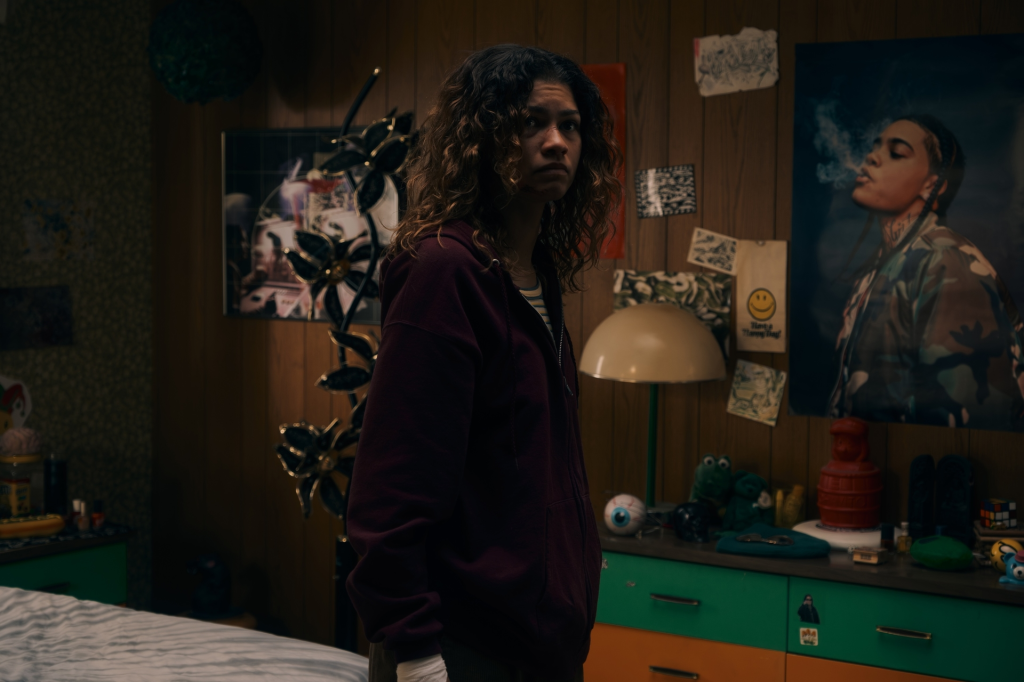 Rue in her bedroom, wearing a maroon hoodie and looking over her shoulder.