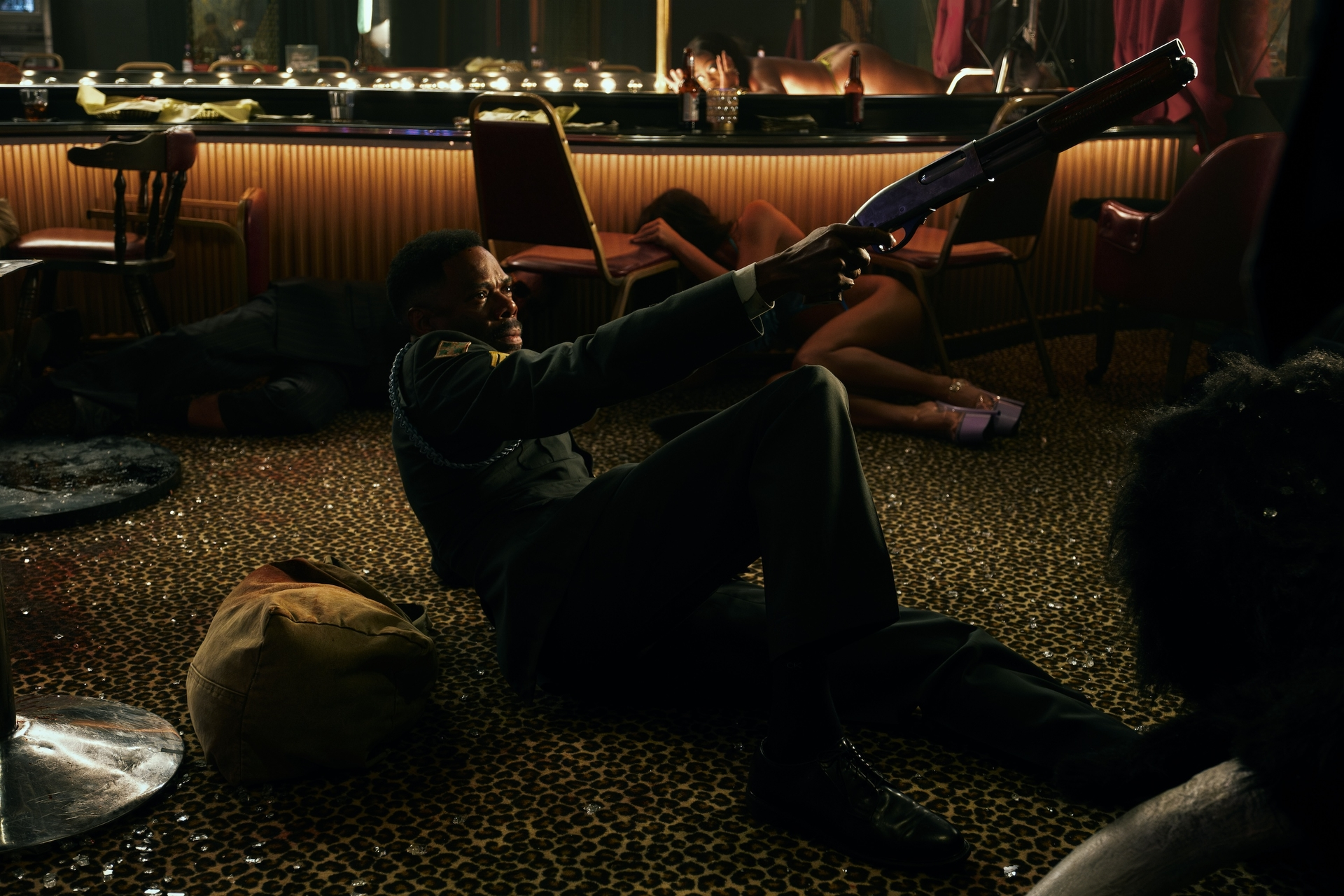 A man in a military uniform lies on a leopard-print floor, aiming a shotgun at someone off-camera, with other people lying on the floor in the background.