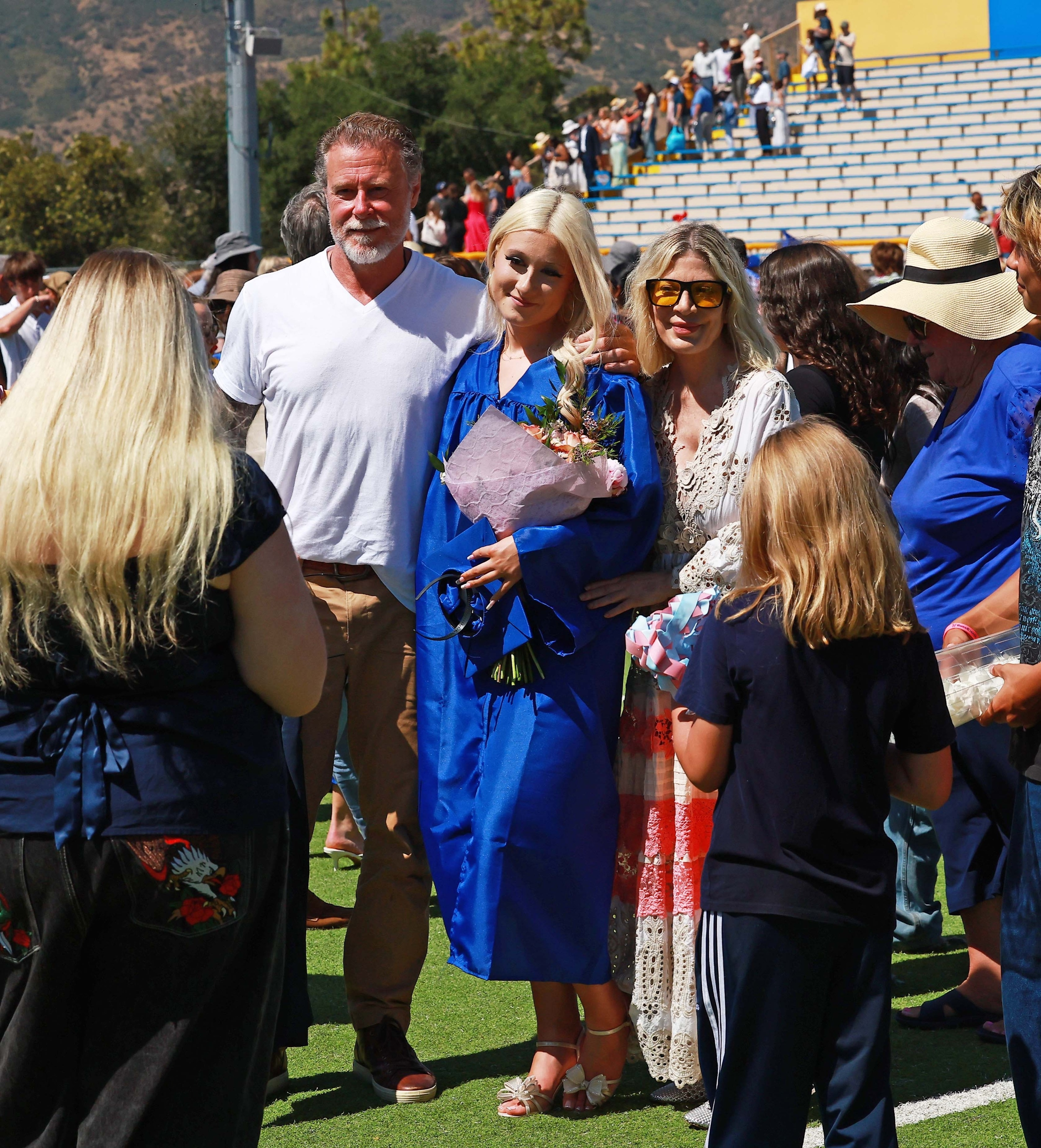 Dean McDermott, his daughter Stella in a blue graduation gown, and Tori Spelling in a white boho-style dress stand on a grassy field.