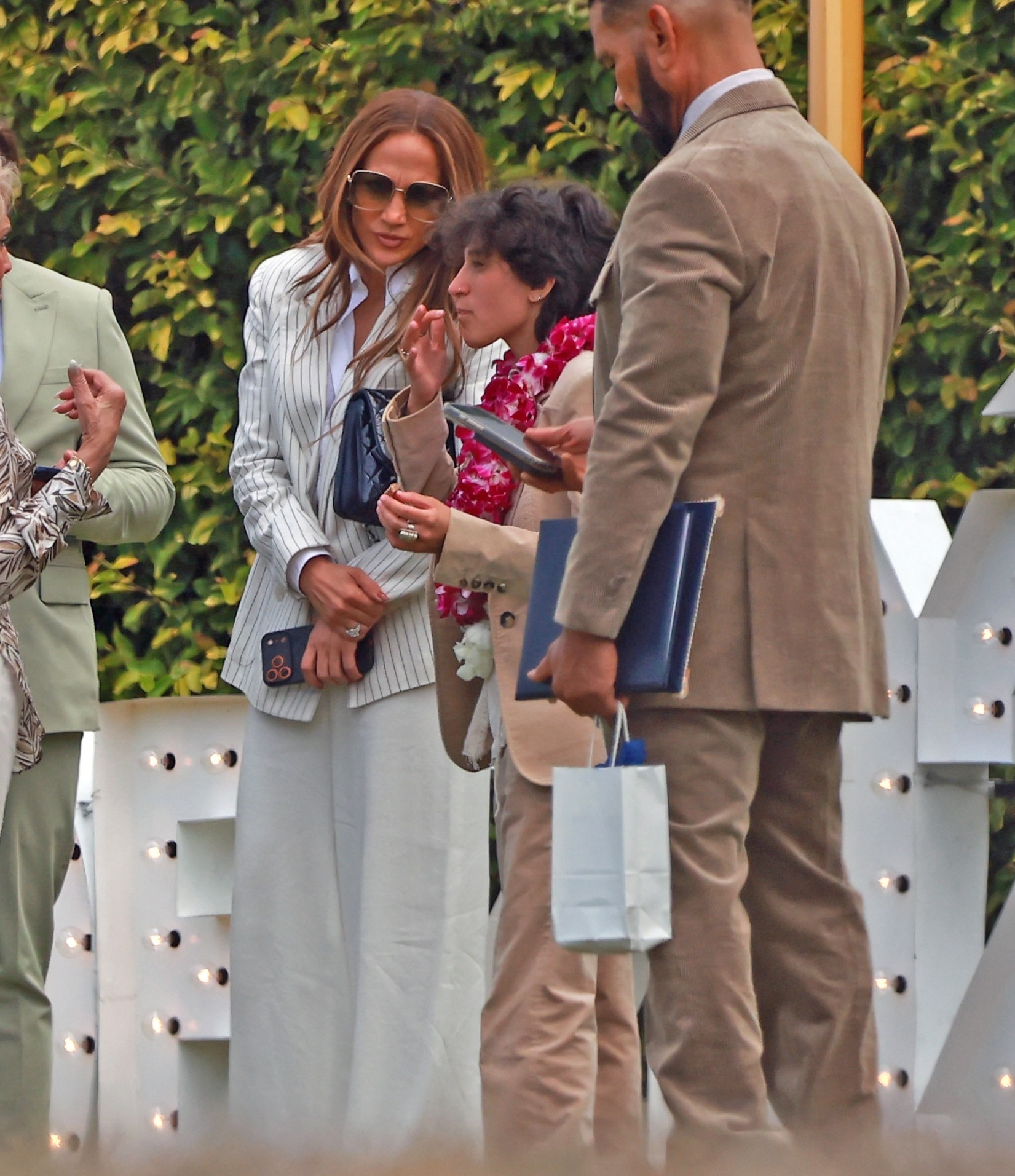 Jennifer Lopez, in sunglasses and a white striped outfit, with Emme Maribel Muñiz wearing a lei, at Emme's high school graduation.