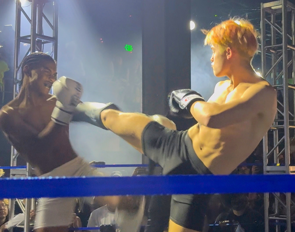 Knox Jolie-Pitt in an exhibition fight, delivering a Muay Thai kick to his opponent in the ring.