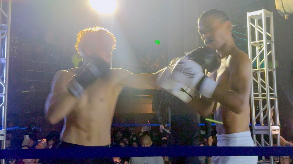 Knox Jolie-Pitt in a boxing ring, wearing white gloves, engaged in an exhibition fight.