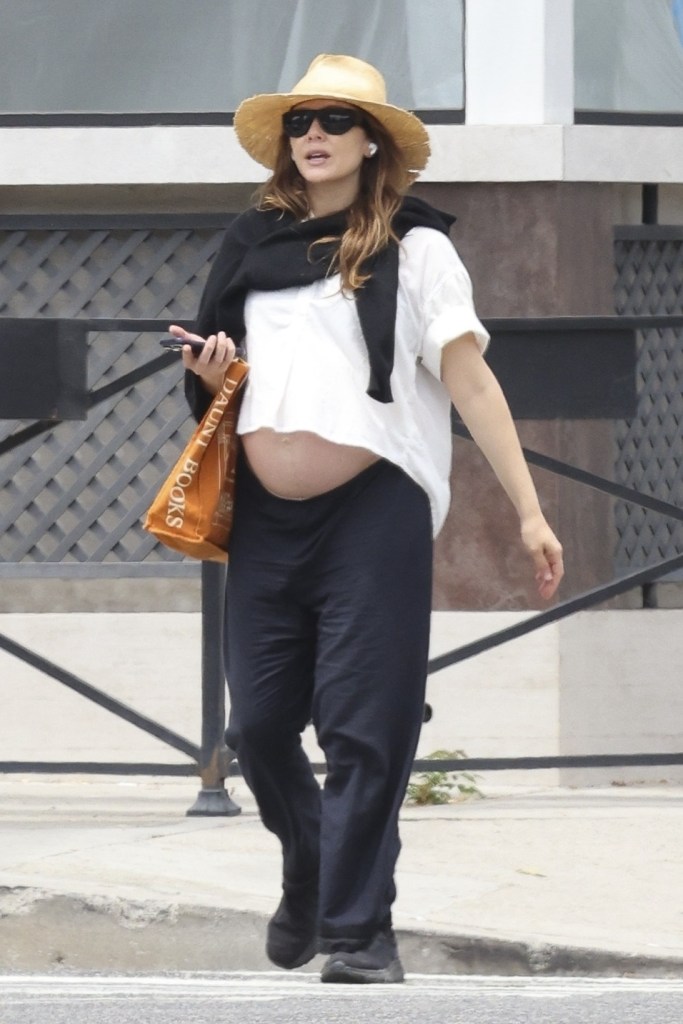 Pregnant Elizabeth Olsen walks outdoors in a white shirt, showing her bare belly, black pants, straw hat, and sunglasses.