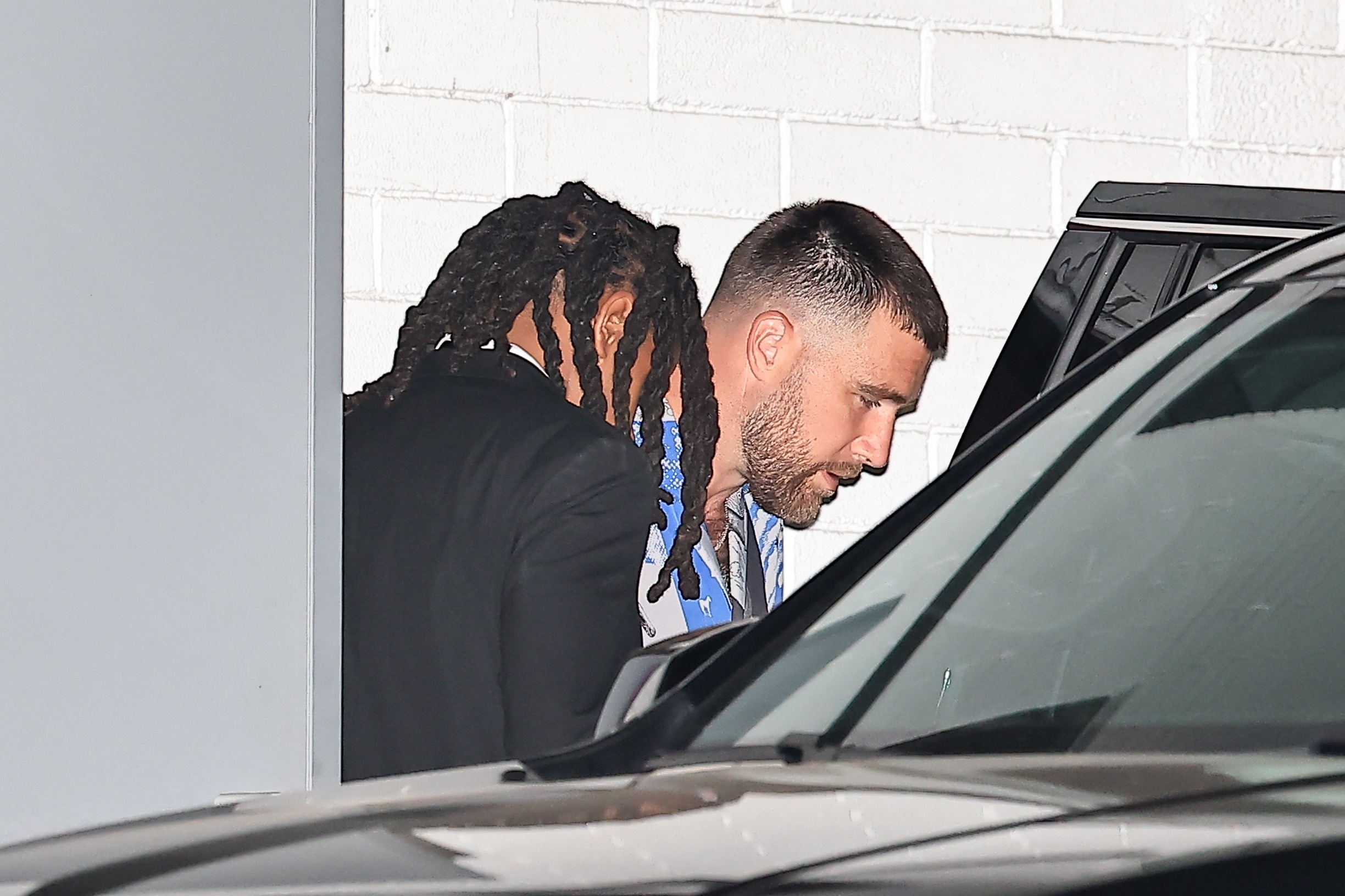 Travis Kelce, with a short beard and faded haircut, looks downward while exiting a car, partially obscured by a person with dreadlocks and a white brick wall in the background.
