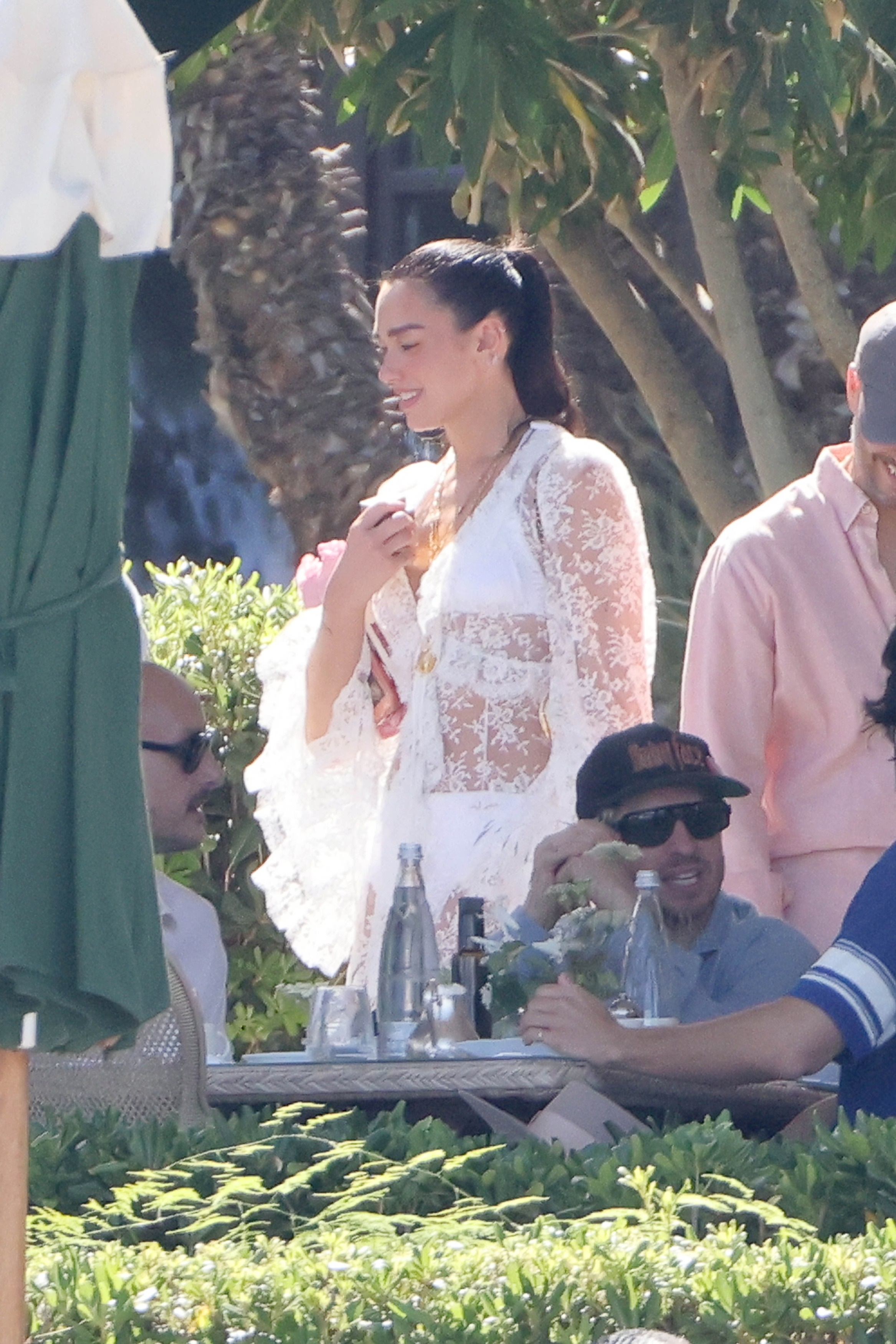 Dua Lipa wearing a sheer white lace dress and golden necklaces at a post-wedding gathering.