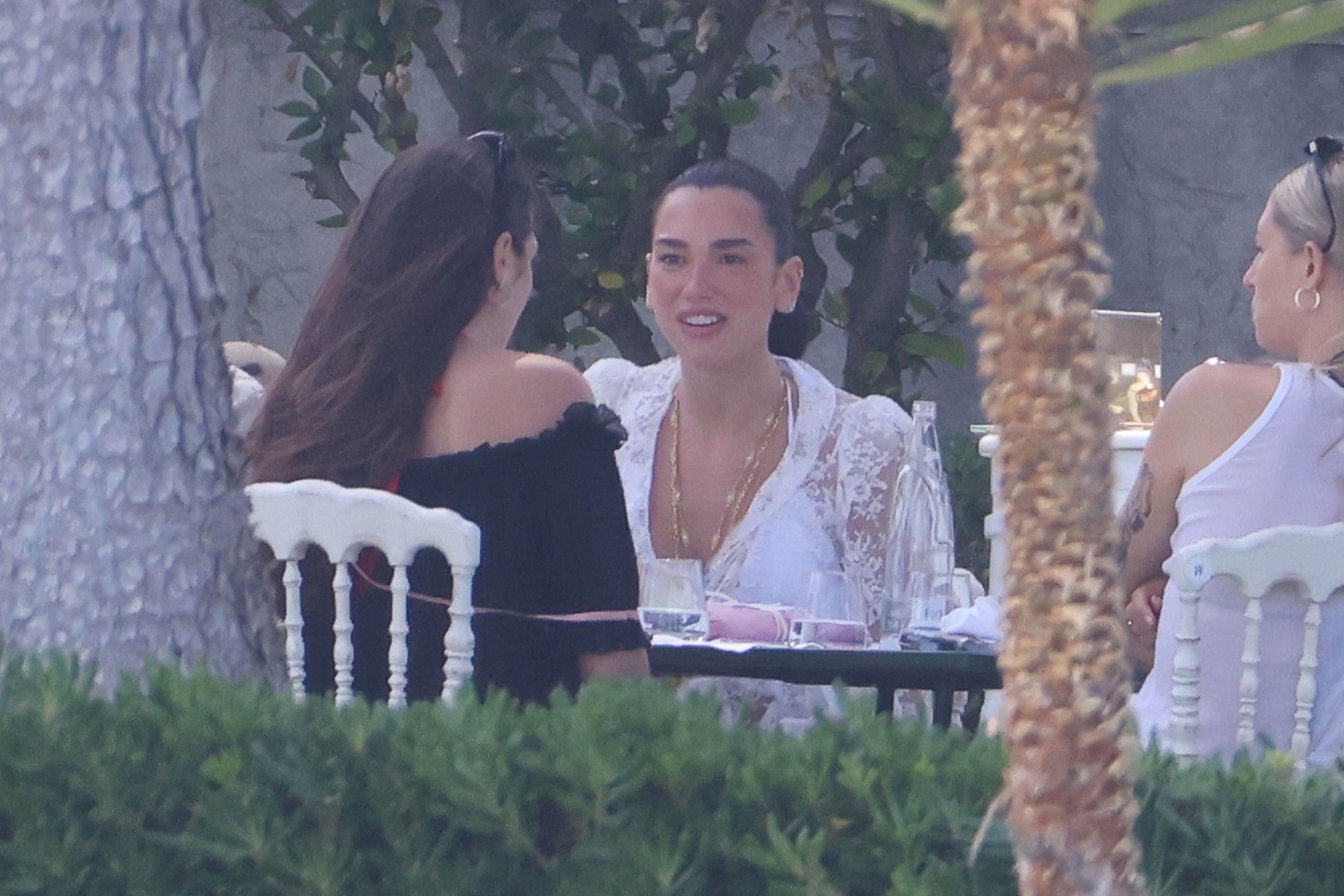 Dua Lipa wearing a sheer white lace top with floral embroidery and gold necklaces at a post-wedding gathering.