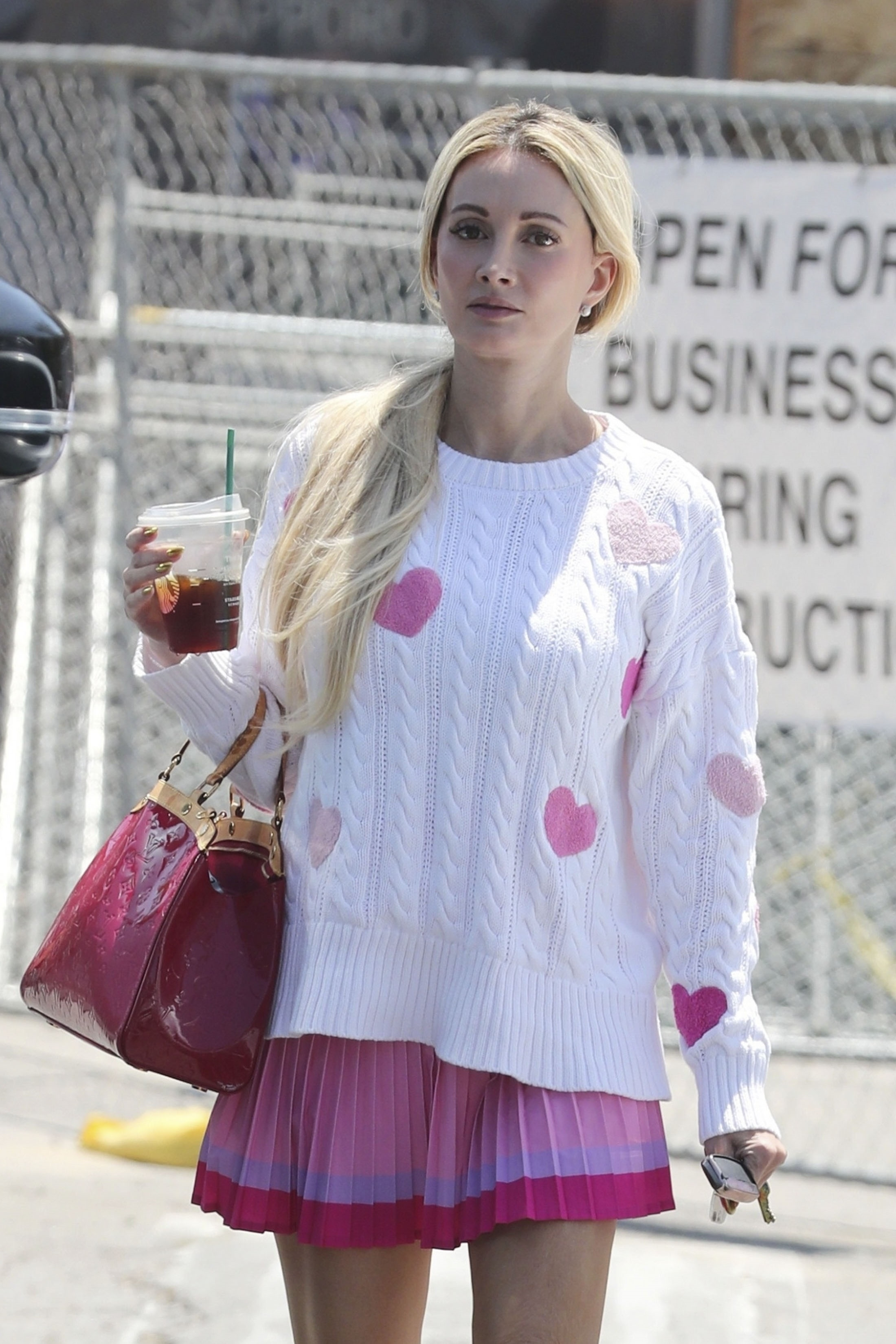 Holly Madison wearing a white cable-knit sweater with pink hearts, a layered pink mini skirt, and carrying a Starbucks drink and a red handbag.