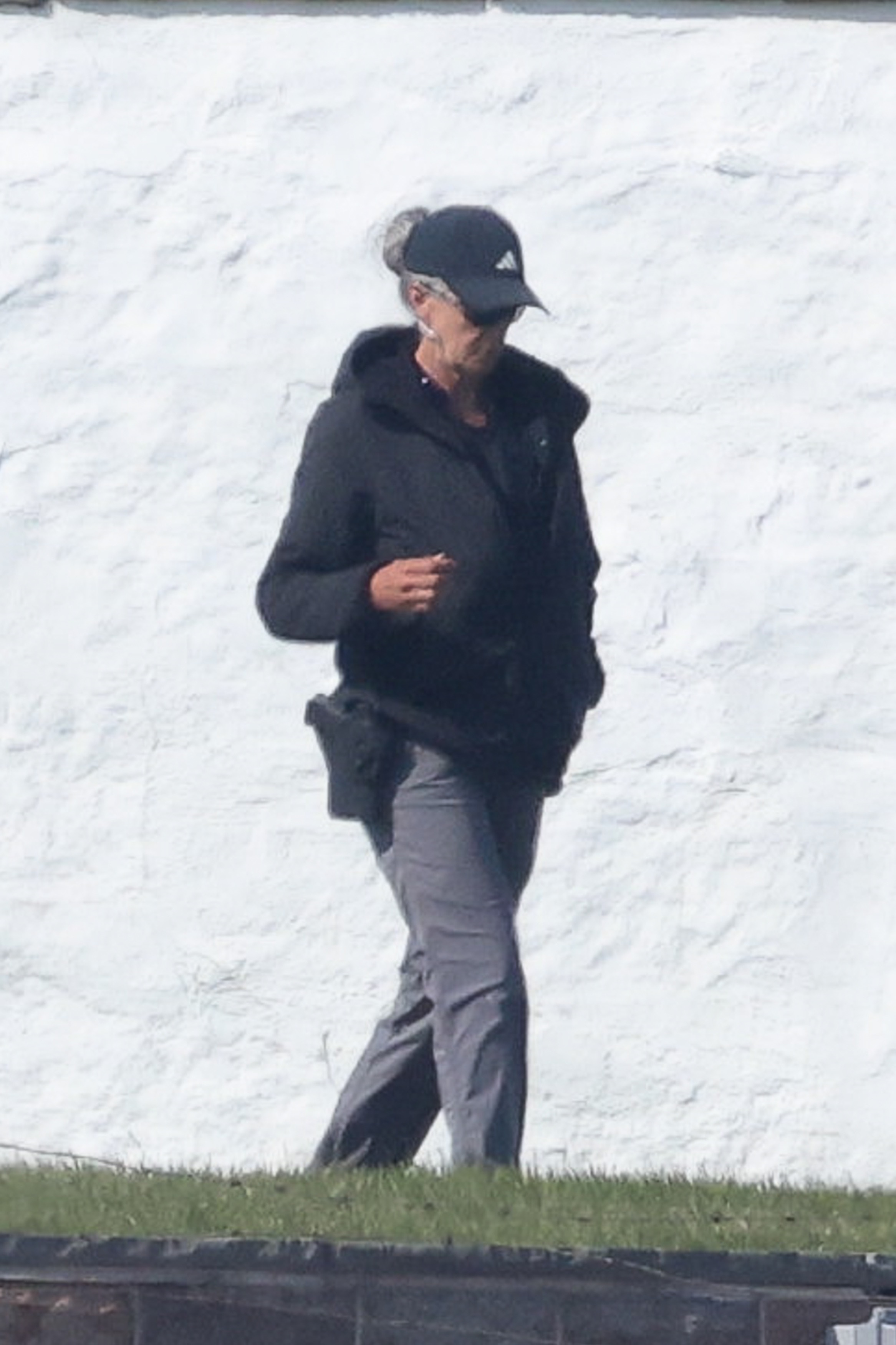 A security guard, wearing a black cap, black jacket, and gray pants, with what appears to be a gun on her belt, walking in front of a white building.