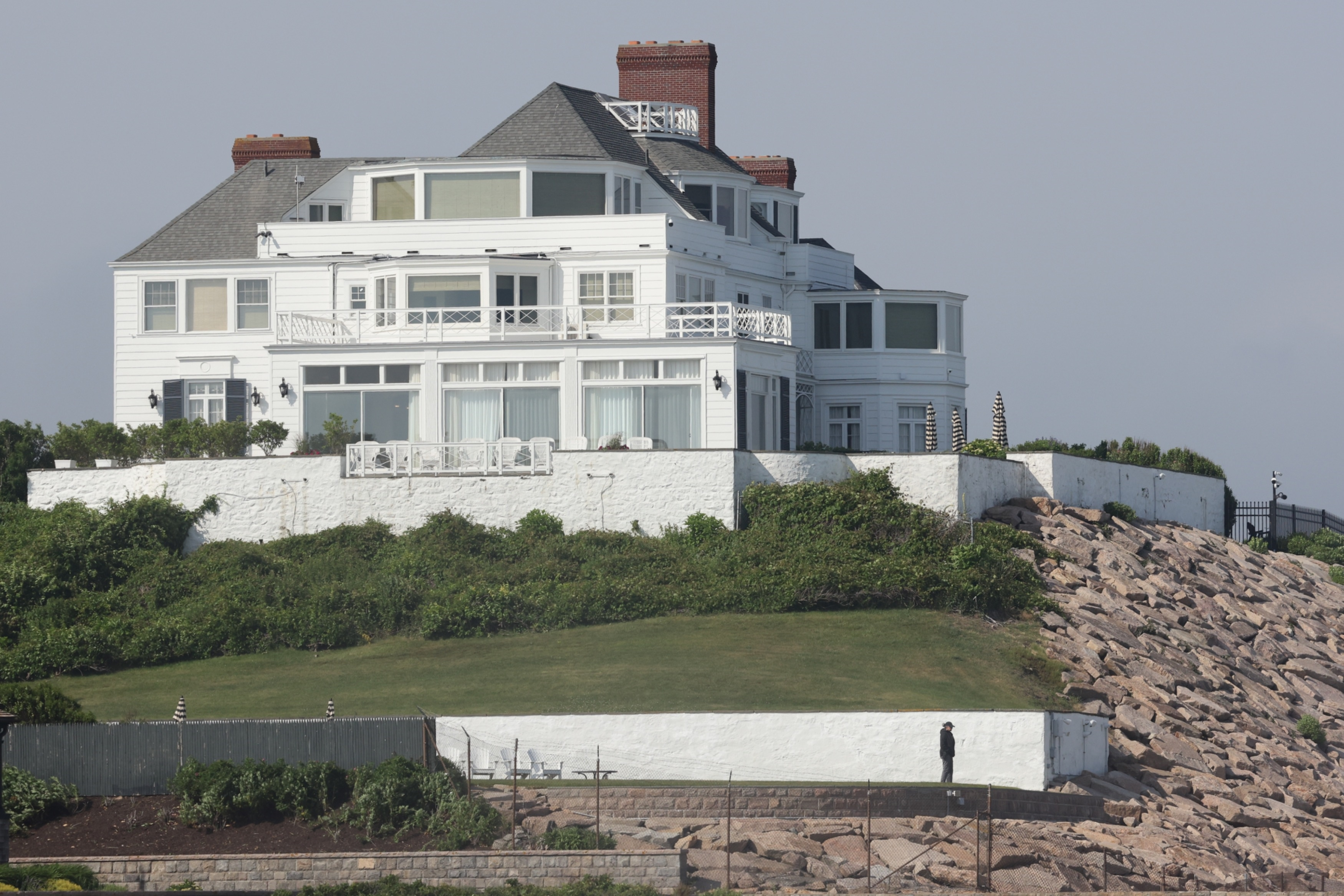 A large white house with many windows sits on a grassy hill overlooking a rocky embankment where a security guard stands.