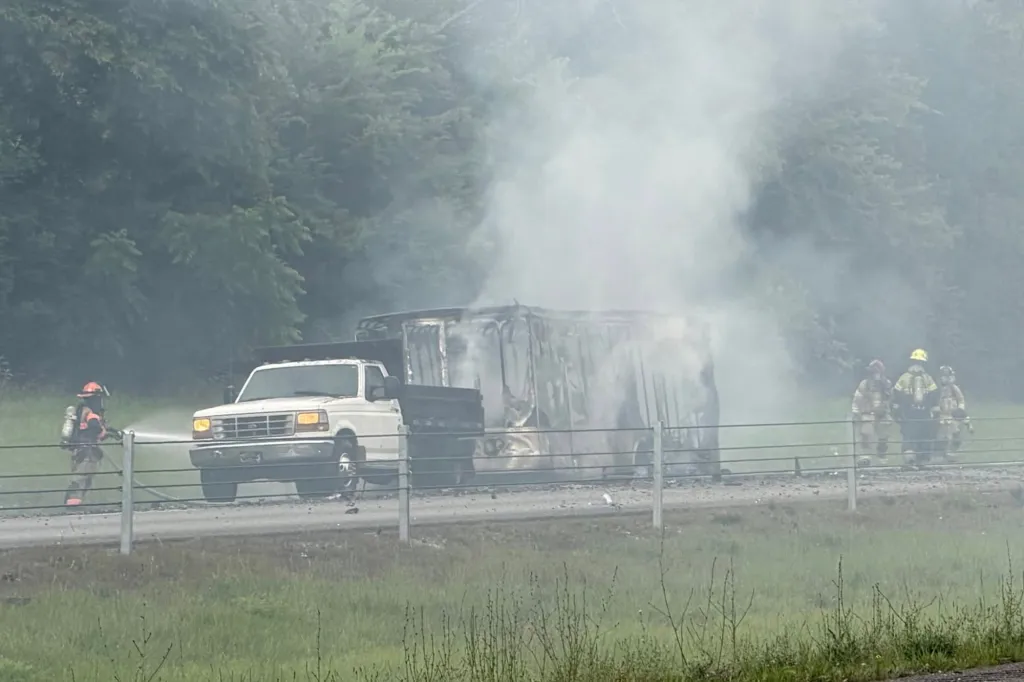Firefighters spraying water on a truck that has caught fire and is smoking heavily on a road next to a grassy area and trees.