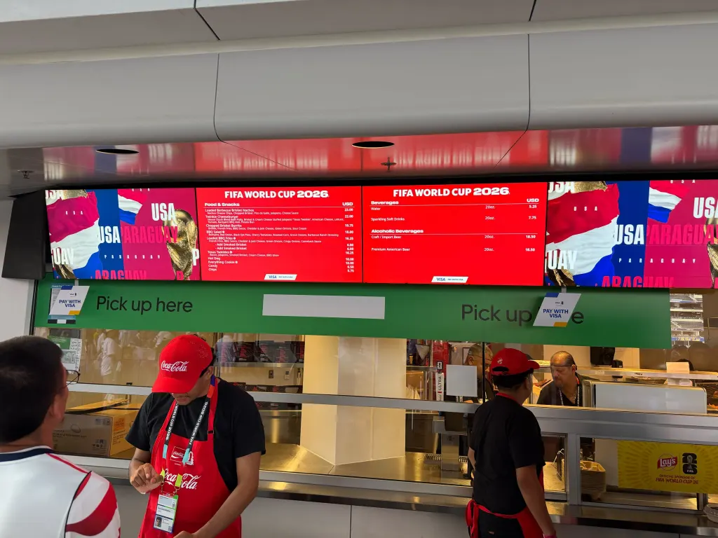 A digital menu board at a FIFA World Cup 2026 concession stand displays high prices for food and drinks, with staff in Coca-Cola aprons below.