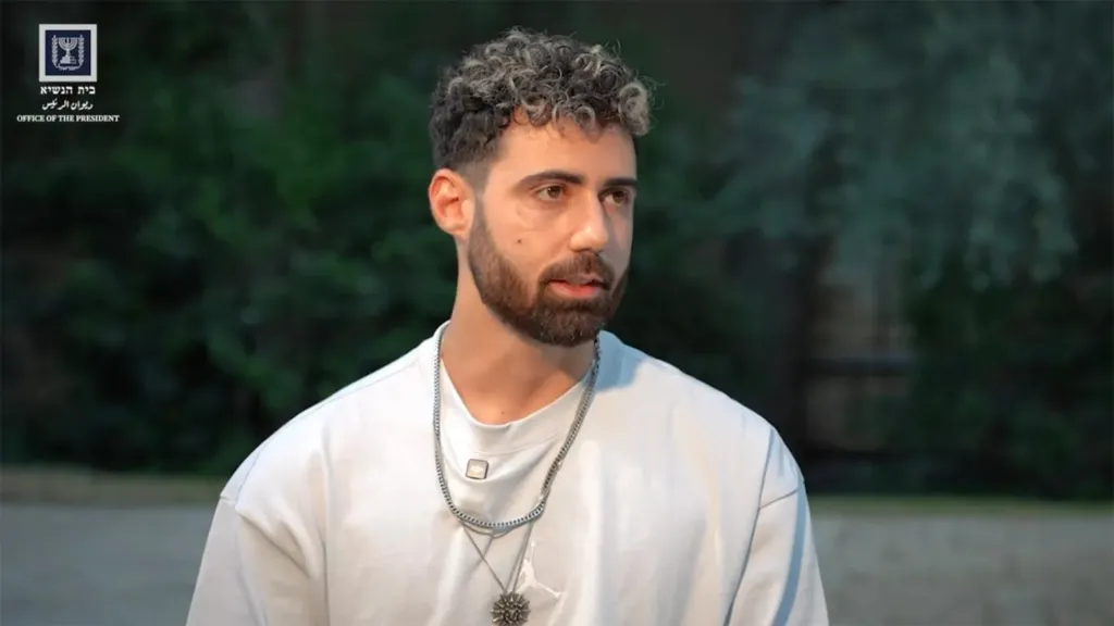 Former Gaza hostage Guy Gilboa-Dalal with curly hair and a beard, wearing a white shirt and two necklaces.