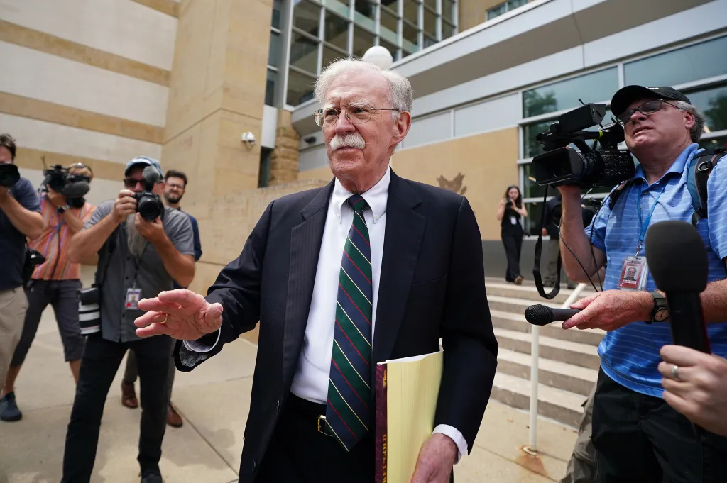 John Bolton, wearing a suit and striped tie, departing a courthouse surrounded by reporters and photographers.