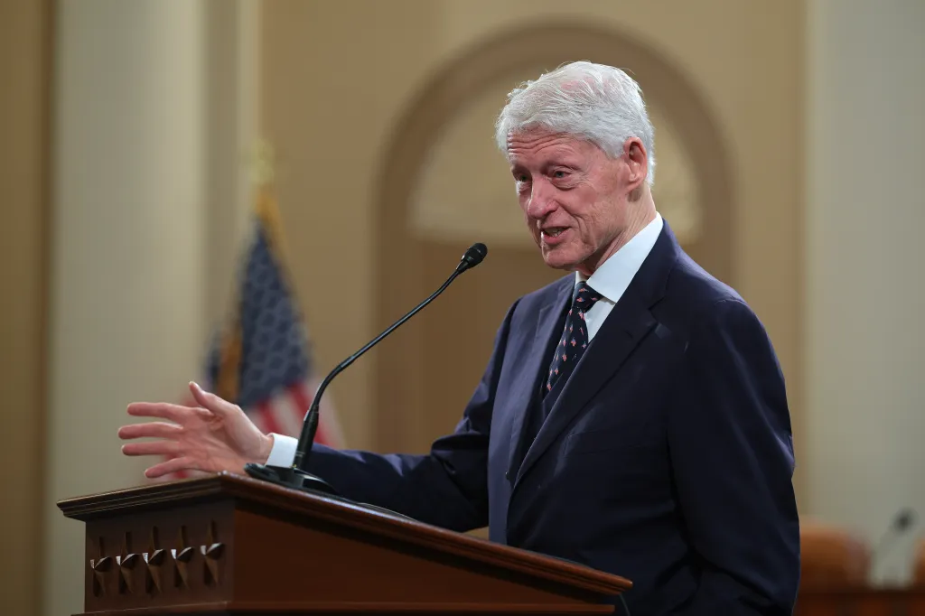 Former U.S. President Bill Clinton speaking at a podium with an American flag in the background.