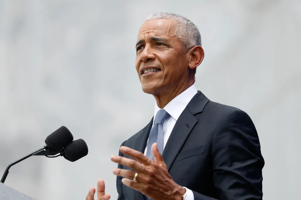 Barack Obama speaking at a podium with two microphones, gesturing with his hands.