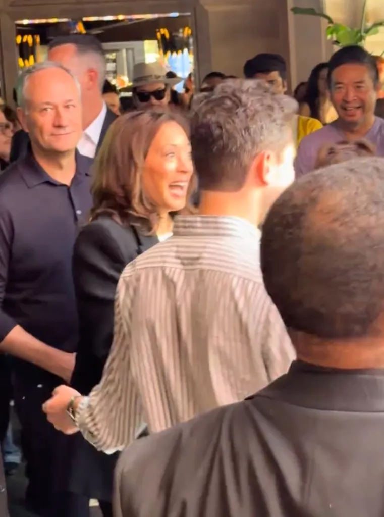 Kamala Harris laughing with a crowd in a bar with her husband, Doug Emhoff, looking on.