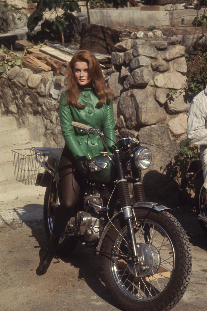 Full-length portrait of Ann-Margret in a green leather jacket and black tights sitting on a motorcycle.