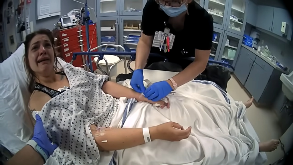 Woman in a hospital bed crying while a medical professional wearing blue gloves attends to her arm.