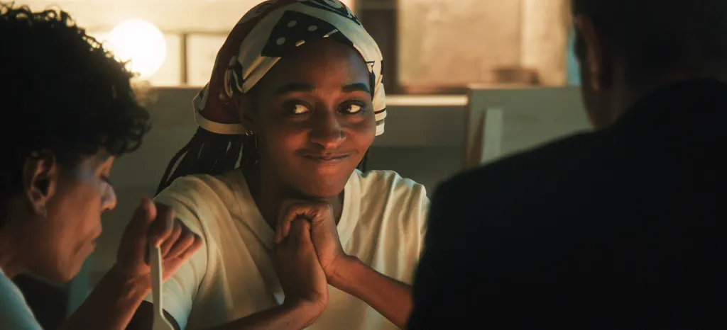 Ayo Edebiri as Sydney Adamu, wearing a patterned headscarf, smiles while looking to her right, with Liza Colón-Zayas as Tina beside her.