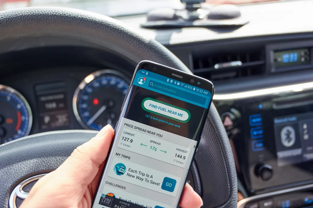A hand holding a Samsung S8 phone displaying the GasBuddy app with fuel prices, inside a car.