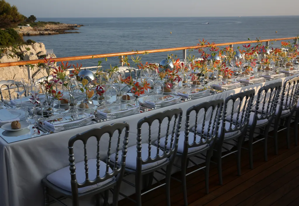Long dining table set for a VIP dinner party, with colorful floral arrangements and the sea in the background.