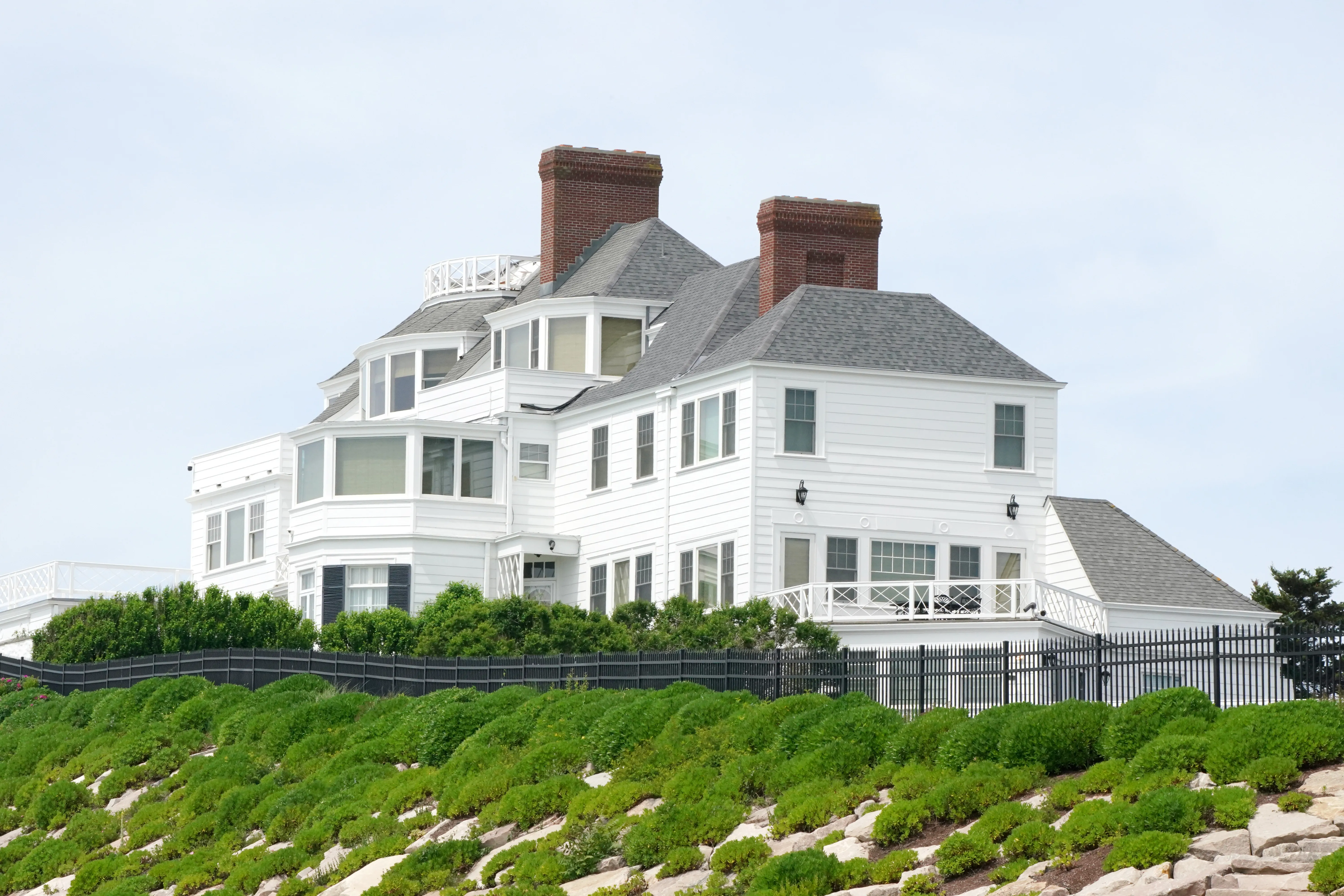 Taylor Swift's Rhode Island home, a large white house with multiple windows and chimneys, visible over a black fence and green landscaping.