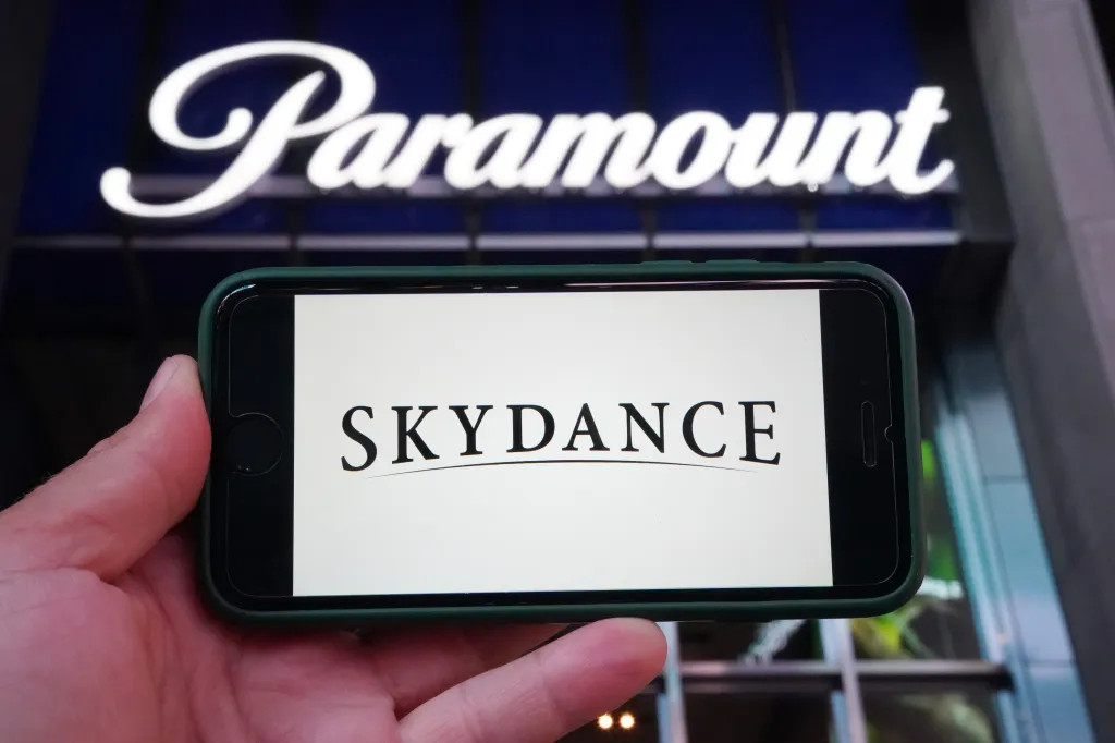 A person's hand holding a phone displaying the Skydance logo, with the illuminated Paramount logo in the background.