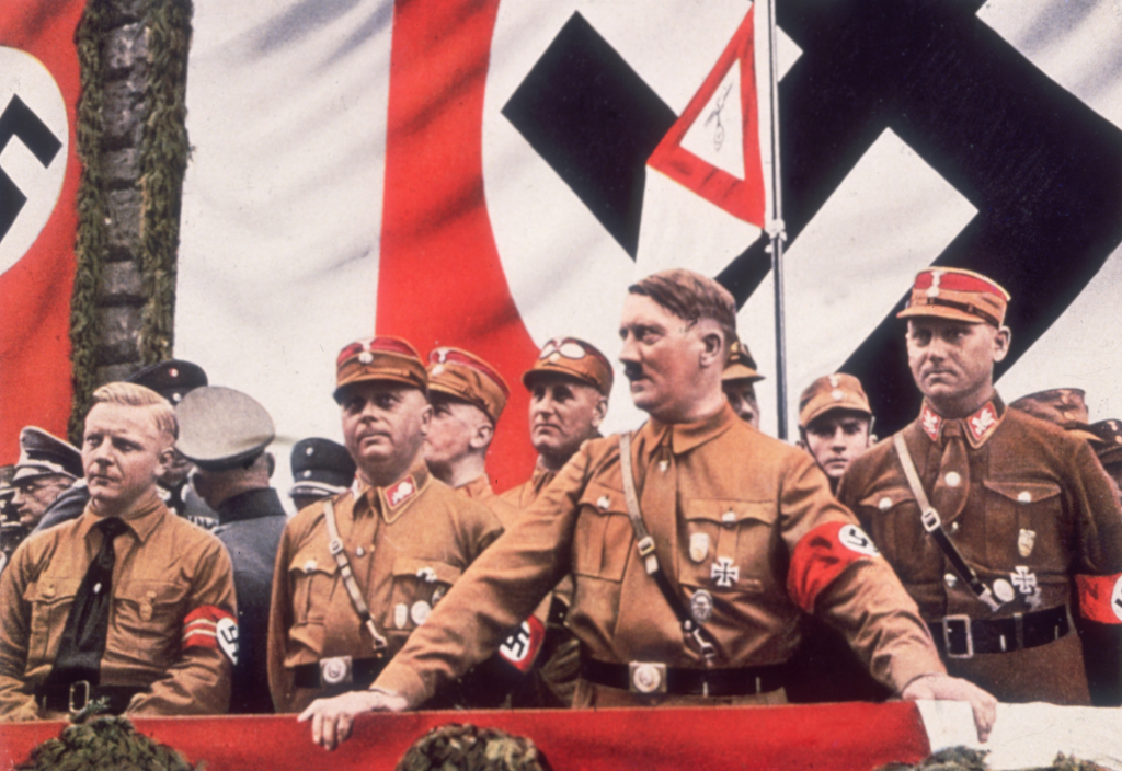 Adolf Hitler addressing a rally in Germany, with several men in uniform beside and behind him, and large Nazi flags in the background.