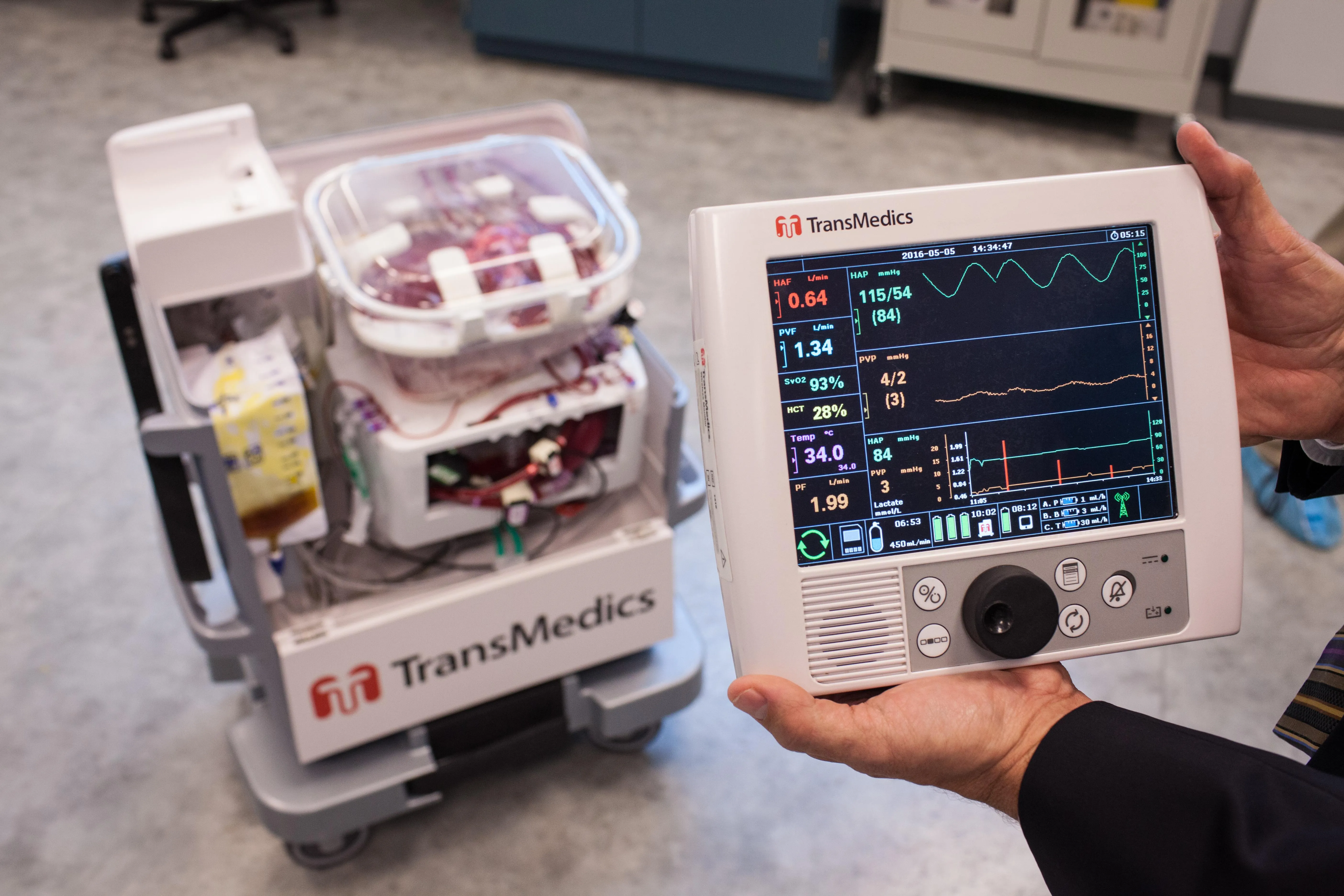 A man’s hands hold an electronic medical device in the foreground while a robotic cart sits in the background