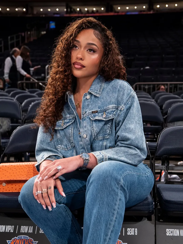 Jordyn Woods sitting on a court-side chair in an empty arena, wearing a denim jacket and jeans.