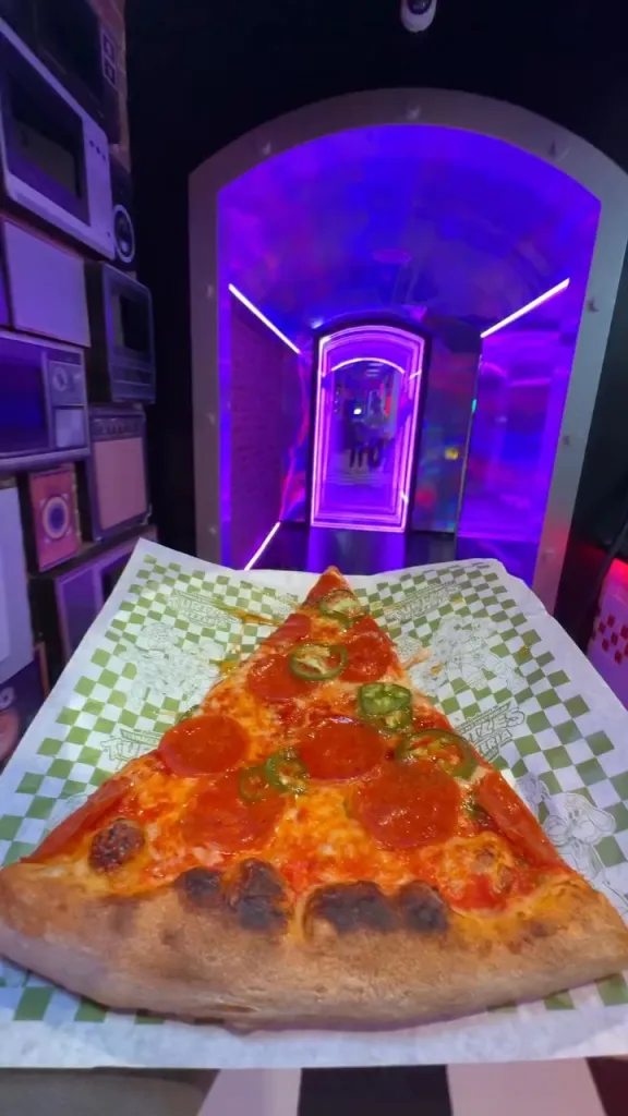 Slice of pepperoni and jalapeño pizza on checkered paper in a purple-lit corridor.