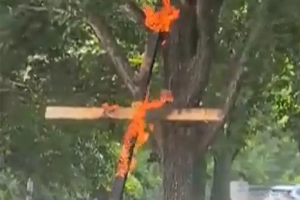 A wooden cross engulfed in bright orange flames leans against a tree.