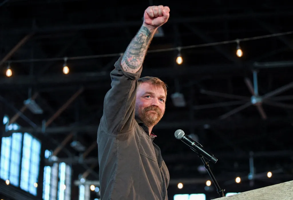 Graham Platner gestures with a raised, tattooed fist while speaking at a campaign rally.