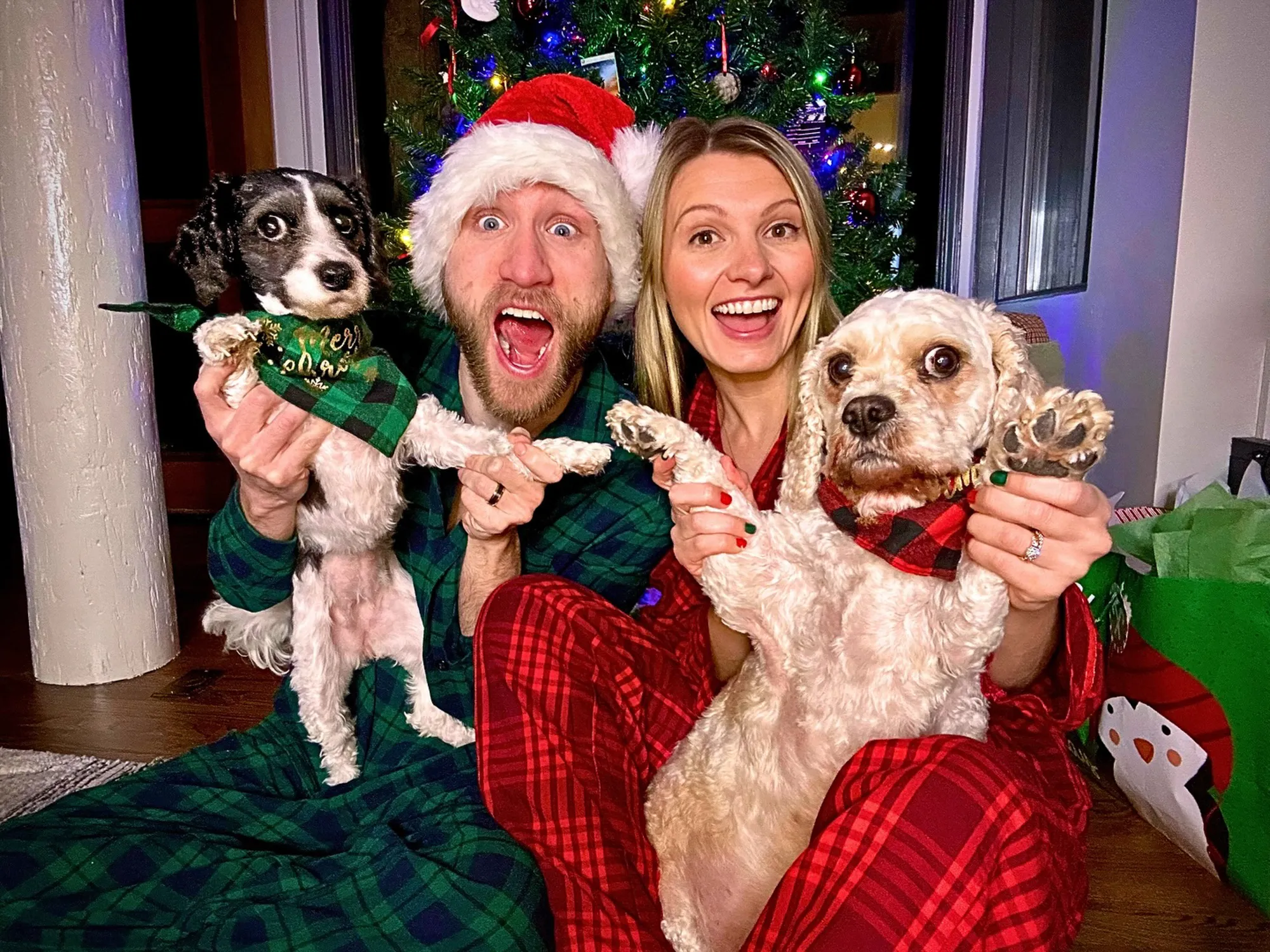 Jesse Ridgway in a Santa hat and his wife with two dogs in Christmas bandanas, all wearing plaid pajamas, in front of a Christmas tree.