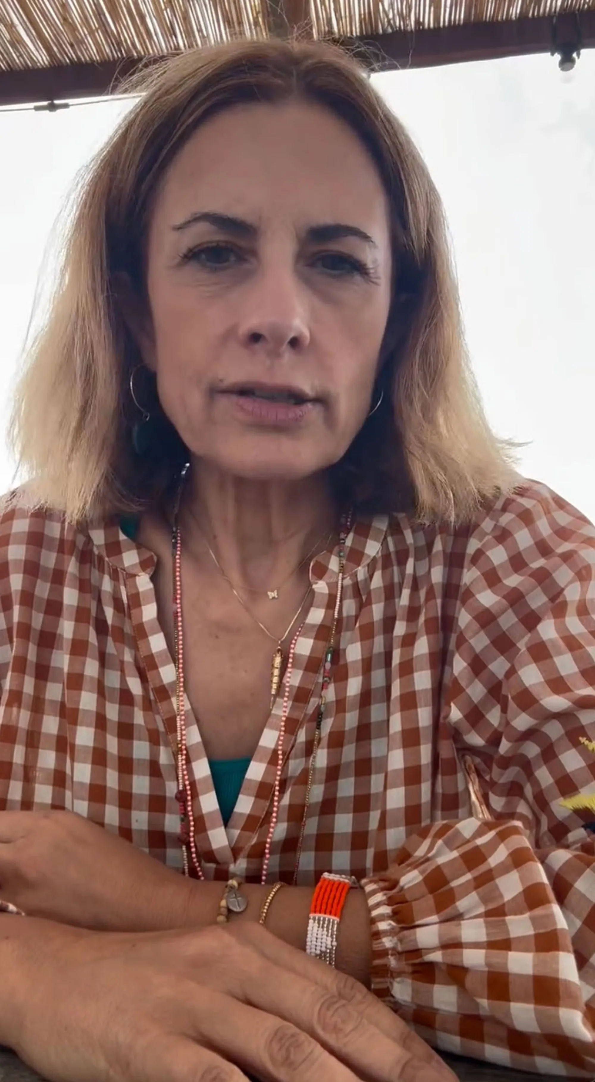 A woman with blonde hair, wearing a brown and white checkered top and multiple necklaces, speaks to the camera.