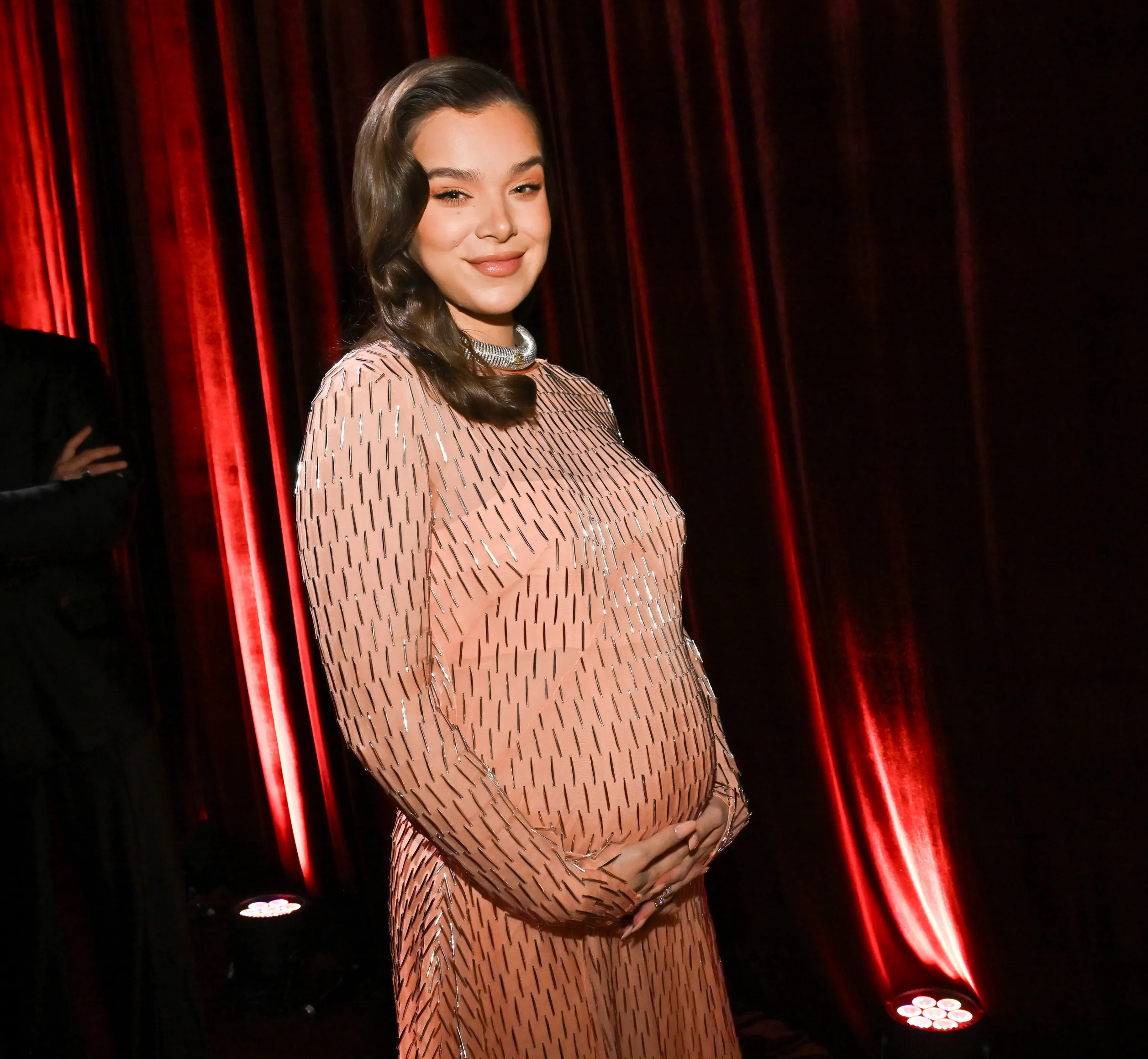 Hailee Steinfeld, in a peach long-sleeved gown, stands on a red carpet.