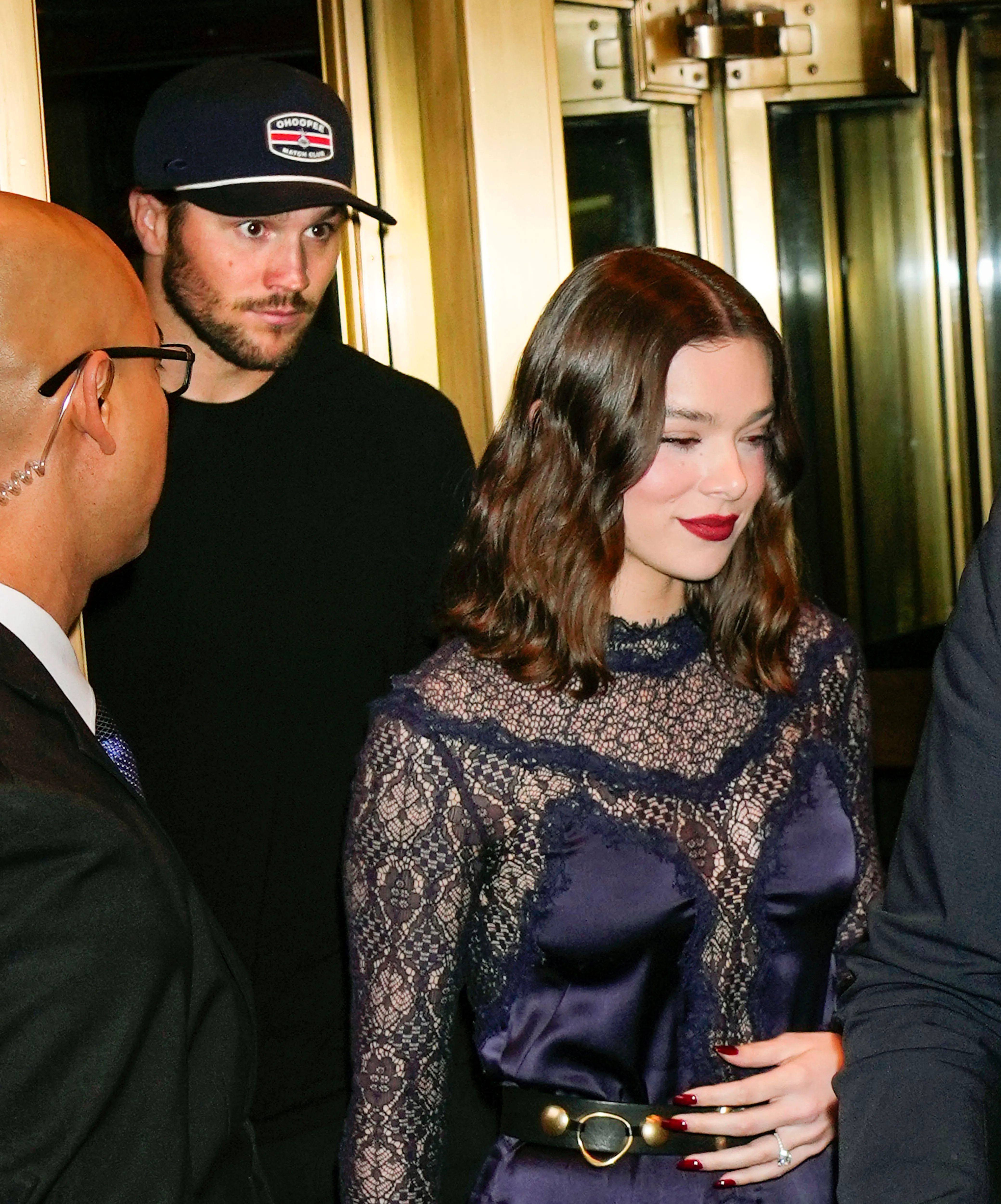 Hailee Steinfeld wearing a purple lace top and Josh Allen in a dark baseball cap, with a security guard in the foreground.